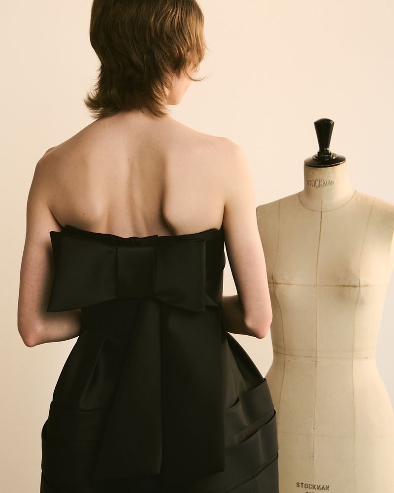 Woman in black strapless dress with bow detail at back standing in front of mannequin. Cream neutral background. Fashion shot.