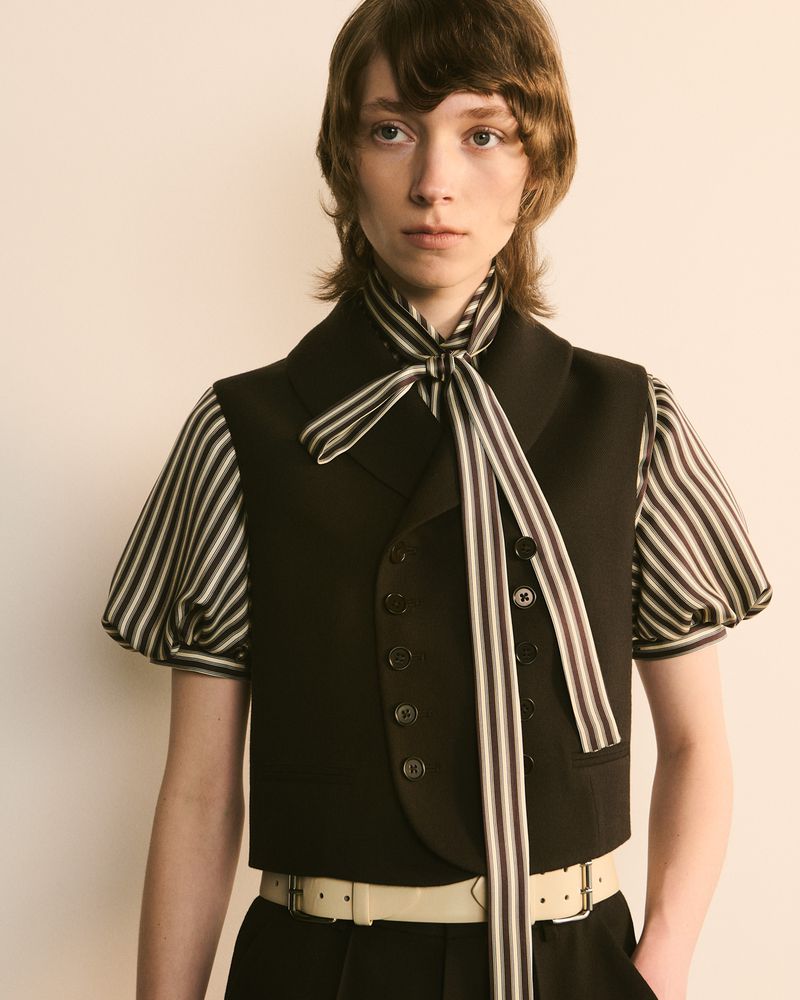 Young adult poses in a cropped black vest, striped collared shirt with bow, and black trousers, set against an off-white backdrop in a fashion shoot.