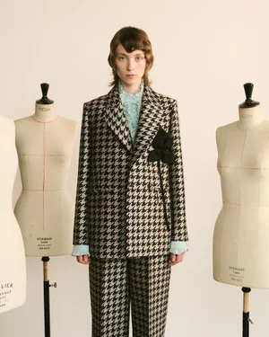 Model wearing a houndstooth suit jacket with a black floral lapel pin standing between two dressmaker mannequins