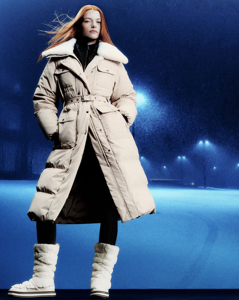 Woman wearing a long, tan puffer coat with a white fur collar and white, furry boots stands outdoors against a blue background with streetlights.