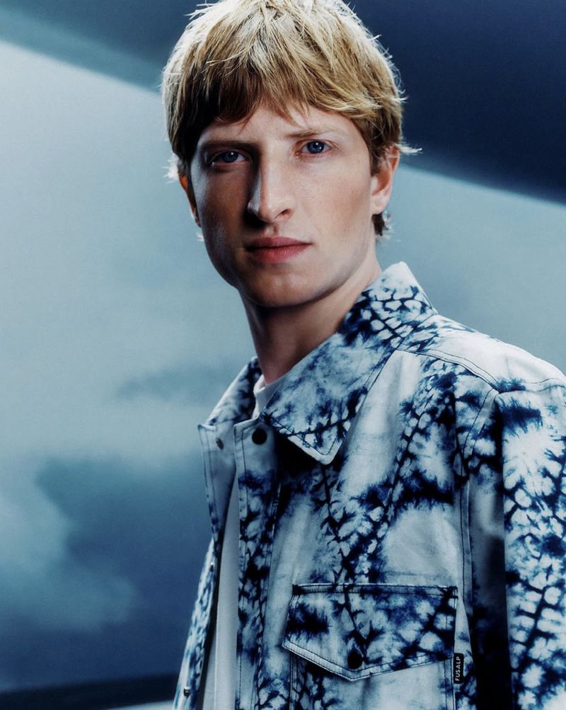 Portrait of a young adult male with blond hair wearing a blue tie-dye jacket in a photo studio with soft lighting.