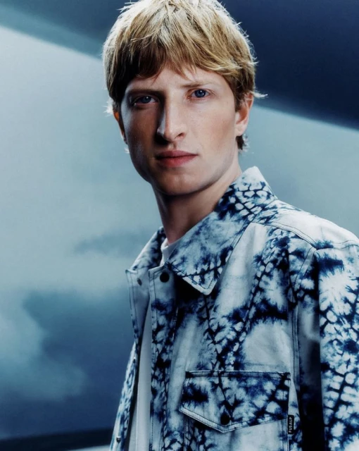 Portrait of a young adult male with blond hair wearing a blue tie-dye jacket in a photo studio with soft lighting.