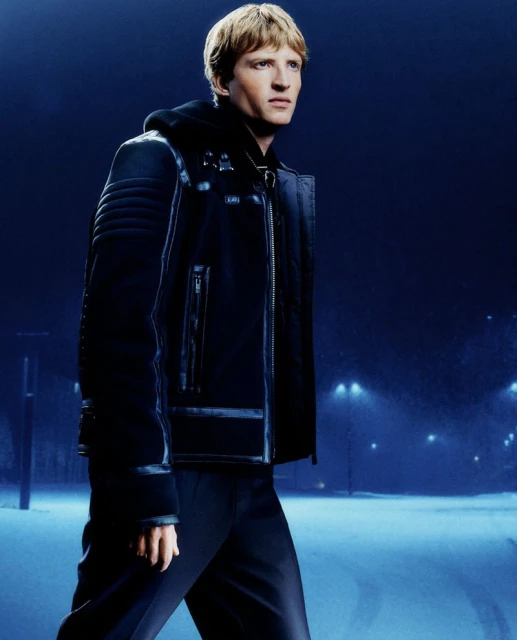 Man in a stylish black winter jacket and dark pants is walking along a snowy path under a dark blue sky with streetlights at night.