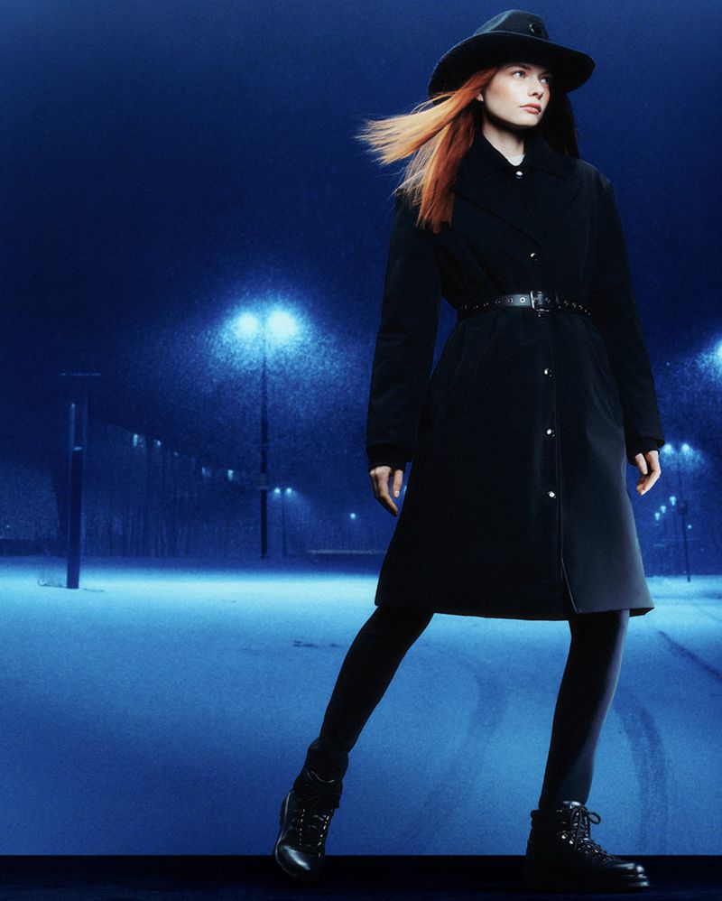 Full-length portrait of a stylish adult female in a dark coat, hat, and boots posing on a snowy urban street at night under streetlights.