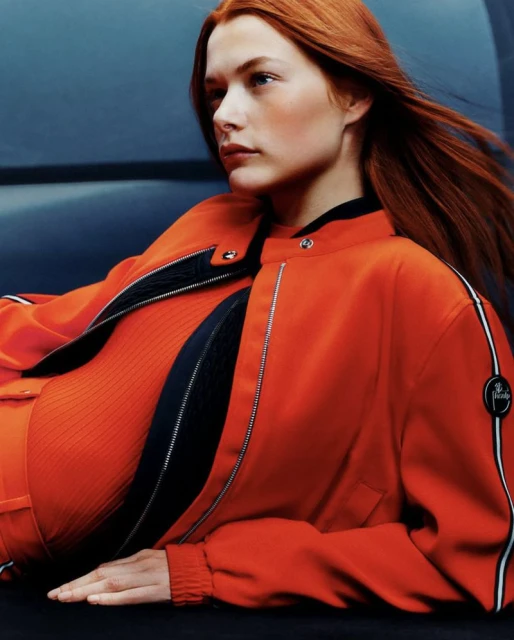 Portrait of a redhead female model wearing an orange bomber jacket and matching top, posing indoors in front of a dark blue background.