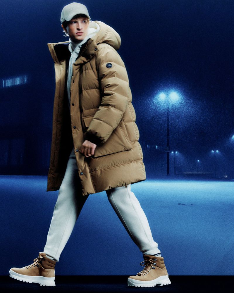 Male fashion model wearing a tan puffer coat, cap, and boots walking in a dark, moody outdoor setting with illuminated streetlights.
