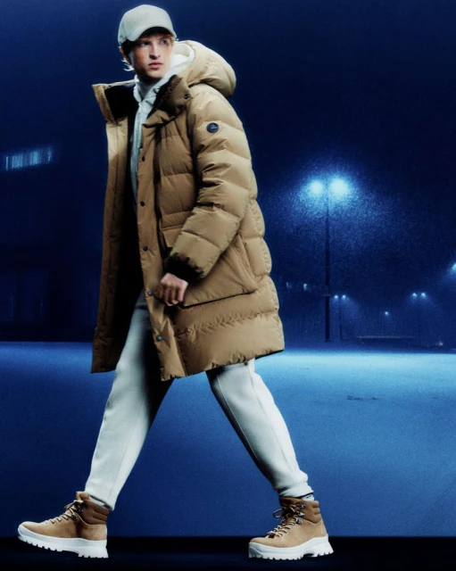 Male fashion model wearing a tan puffer coat, cap, and boots walking in a dark, moody outdoor setting with illuminated streetlights.