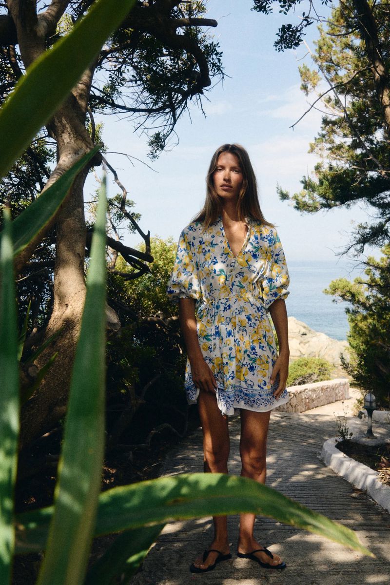 Woman in floral dress stands on stone path surrounded by trees with a glimpse of the ocean in the background on a sunny day.