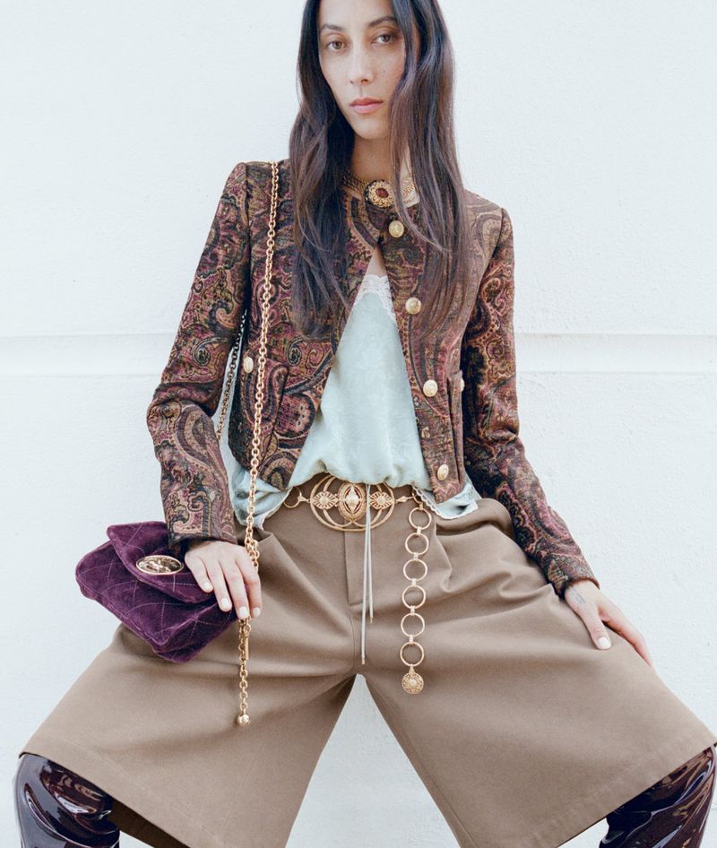 A fashionable adult woman in a patterned jacket poses in a brightly lit studio. She carries a quilted velvet purse with gold chain.