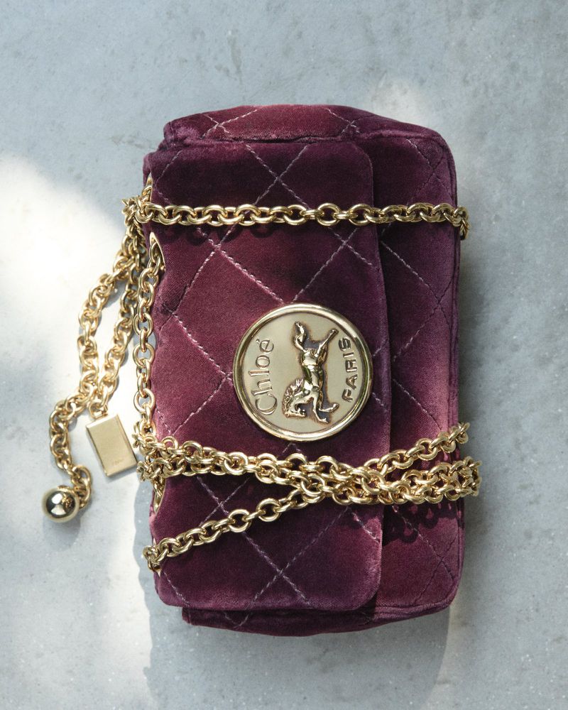 Burgundy velvet quilted purse with gold chain strap and Chloé logo against a gray surface, ideal for fashion or beauty content.