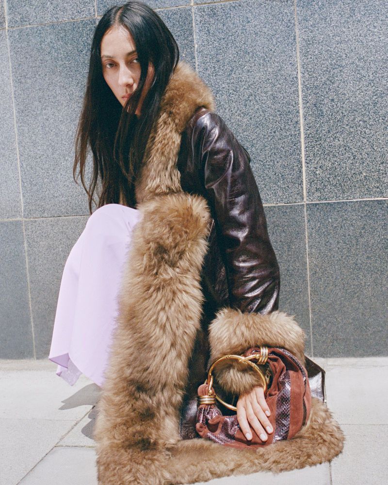 A young adult female model poses in a dark coat with fur trim, a pastel dress, and a brown handbag against a grey wall.
