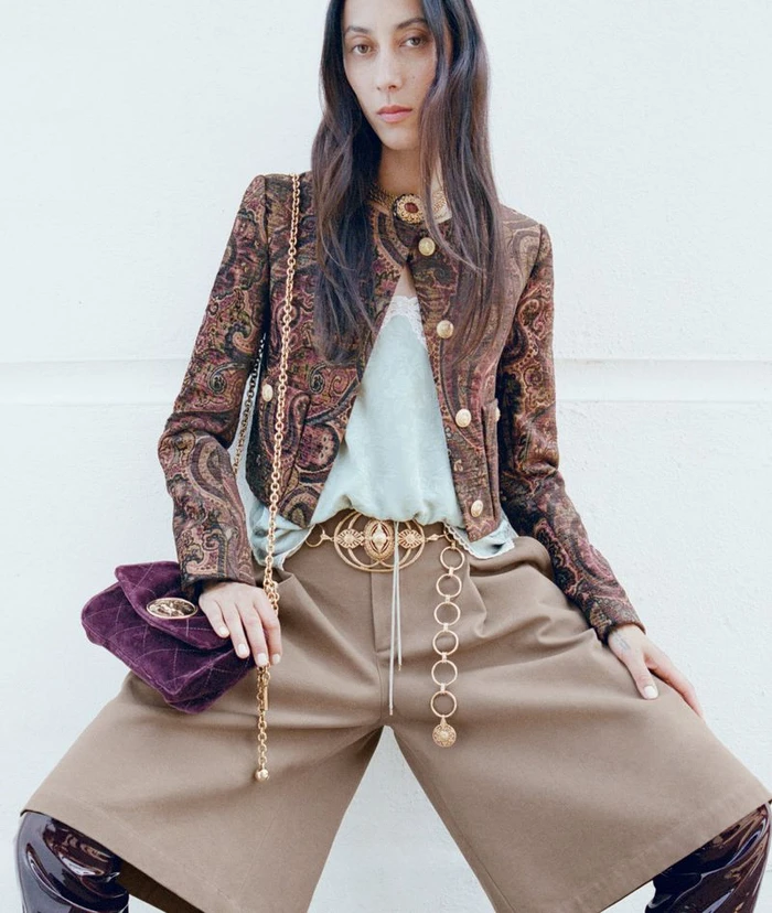 Model wearing a patterned jacket, beige trousers and holding a purple velvet handbag against a plain background