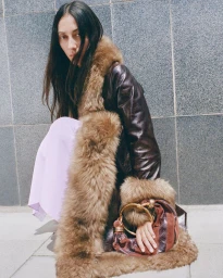Model crouching against a granite wall wearing a dark leather coat with fluffy fur trim and holding a ring-handled handbag