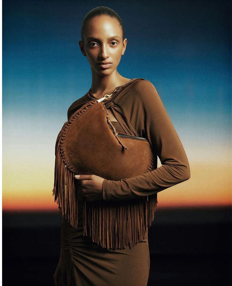 Fashion shot of a woman in a long-sleeved dress holding a fringed brown handbag against a diffused blue and orange background.