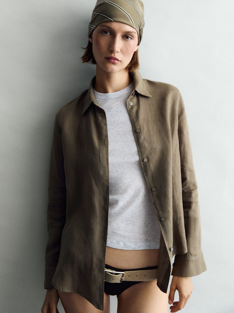 Woman in olive green bandana and matching shirt, shot in studio, leans against a wall. Fashion editorial photograph with neutral tones.