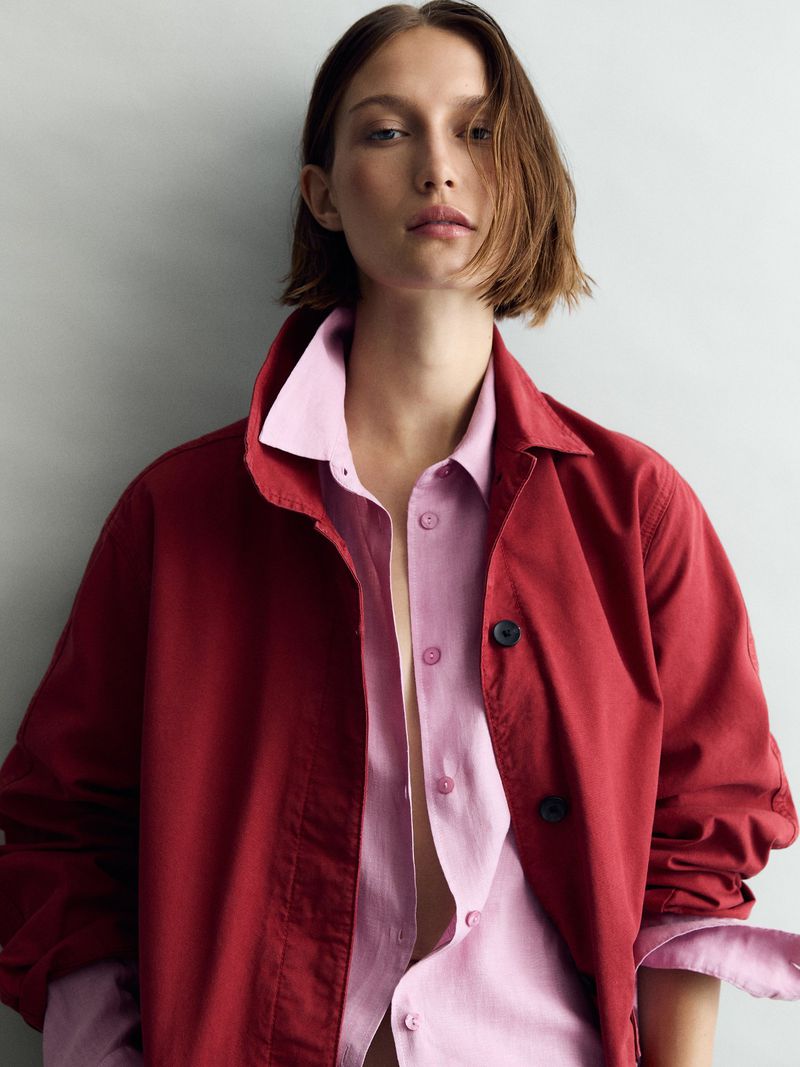 Fashionable young woman in a red jacket and pink button-down shirt gazes serenely at the camera in a studio setting, shallow depth of field.