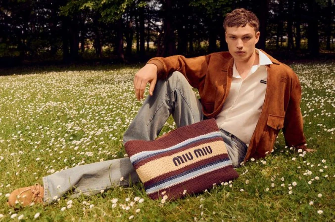 Male model sits in a flower-filled meadow wearing a brown jacket, jeans, and carrying a Miu Miu tote bag in a fashion editorial.