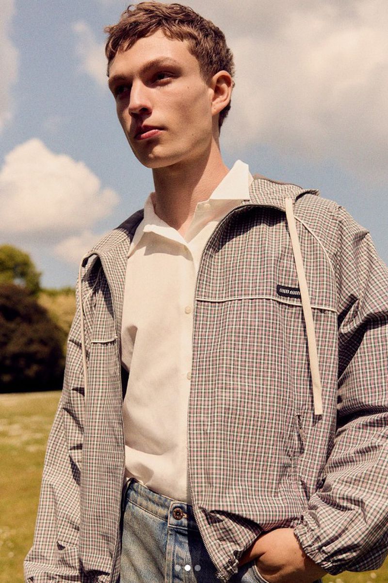Young adult man in checkered jacket and jeans stands in a field of green grass beneath a blue sky with scattered white clouds.