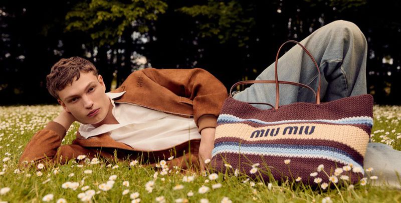 Male fashion model lying on a flower meadow next to a Miu Miu knit handbag in an outdoor setting with trees visible in the background.