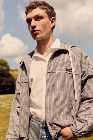 Young man wearing a checkered jacket and white collared shirt outdoors against a cloudy sky.