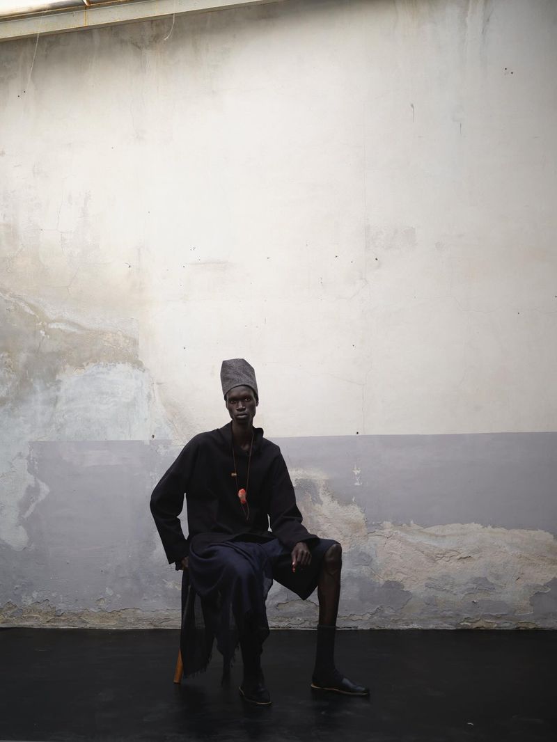 Adult male sits in front of a concrete wall wearing a knit cap, dark shirt, and shorts in a studio with dark floors.
