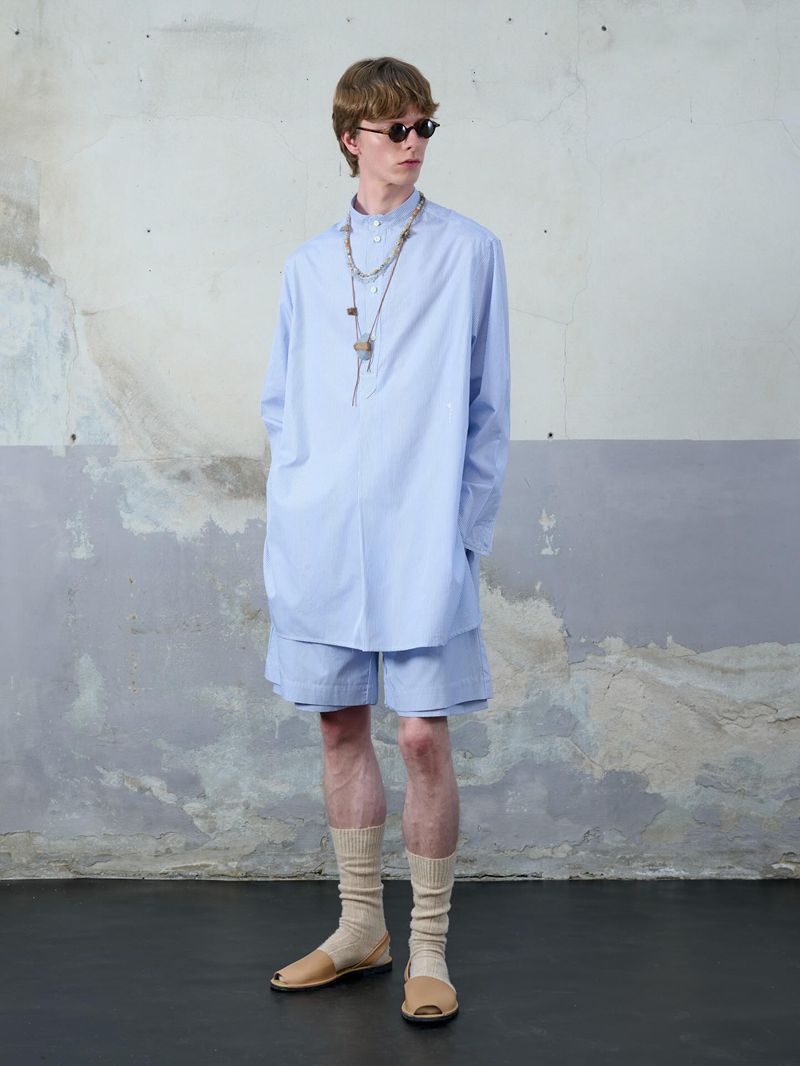 A slender man in a light-blue shirt and shorts stands against a textured wall, showcasing a relaxed, summery fashion aesthetic.