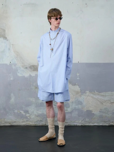 A slender man in a light-blue shirt and shorts stands against a textured wall, showcasing a relaxed, summery fashion aesthetic.