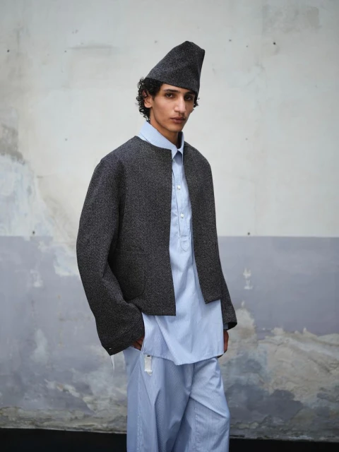 Fashionable young man in blue striped pants and a blue button-up shirt under a cropped grey jacket with a grey pointed hat against a textured wall.
