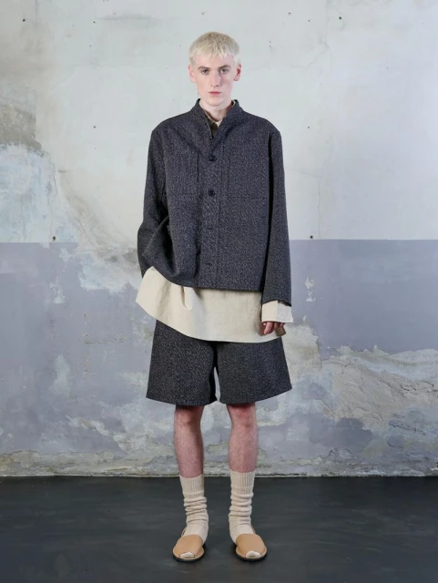 Fashion portrait of a young adult male with platinum hair wearing a gray jacket, beige shirt, and gray shorts in a studio setting.