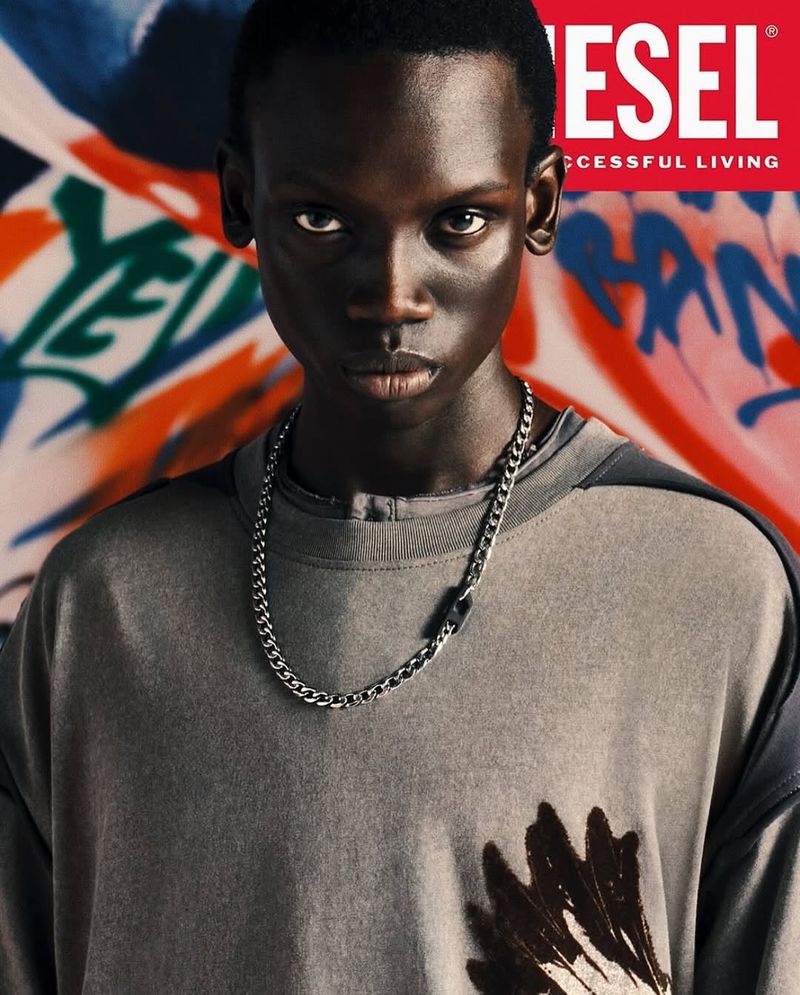 Portrait of a young adult male with dark skin wearing a grey t-shirt and silver chain necklace in front of a graffiti background.