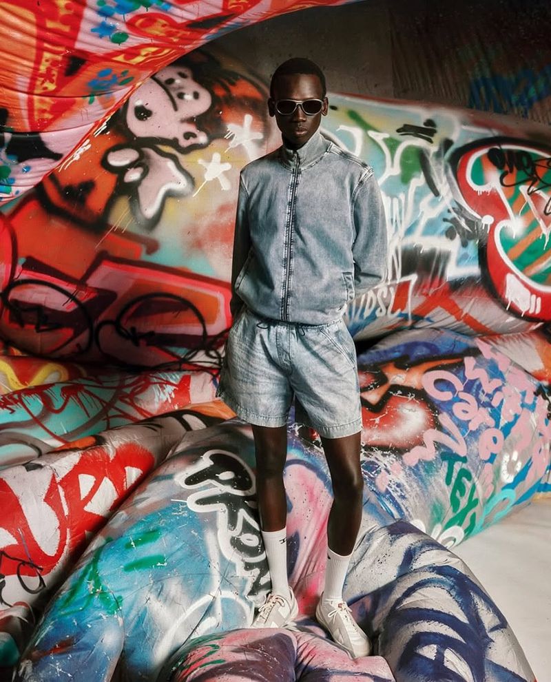 Model Bukwop Kir wearing a light wash denim jacket and shorts with sunglasses against inflatable graffiti art for Diesel.