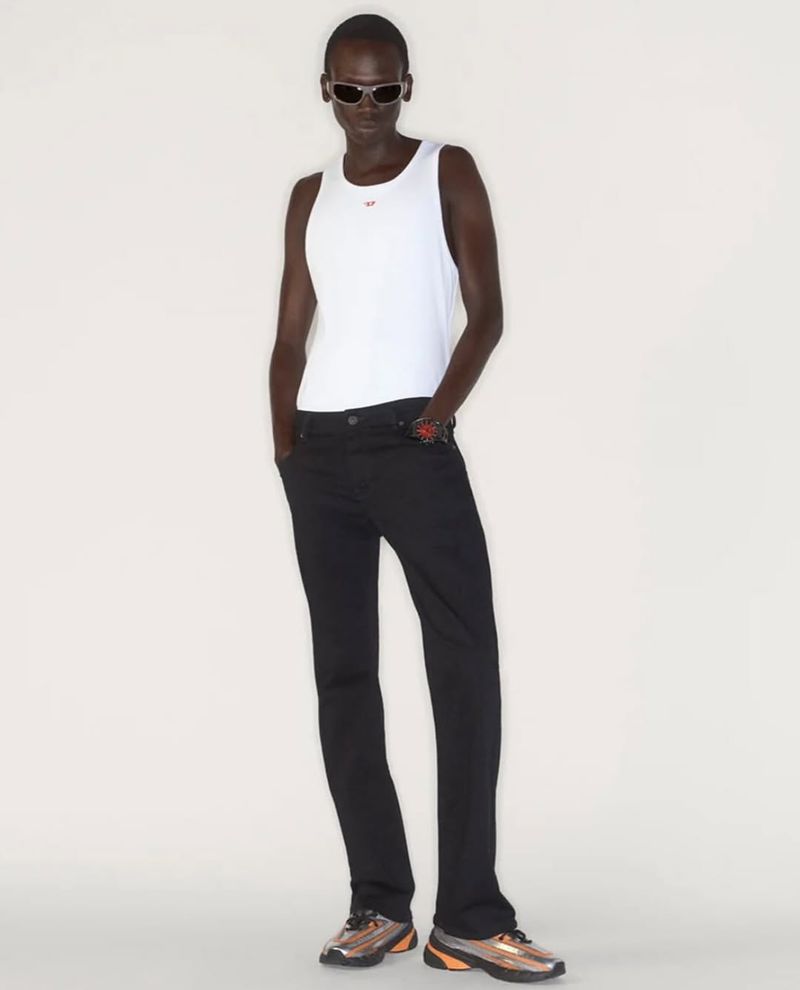 A slender, dark-skinned person in a white tank top and black jeans stands indoors against a neutral backdrop in a full-length fashion shot.