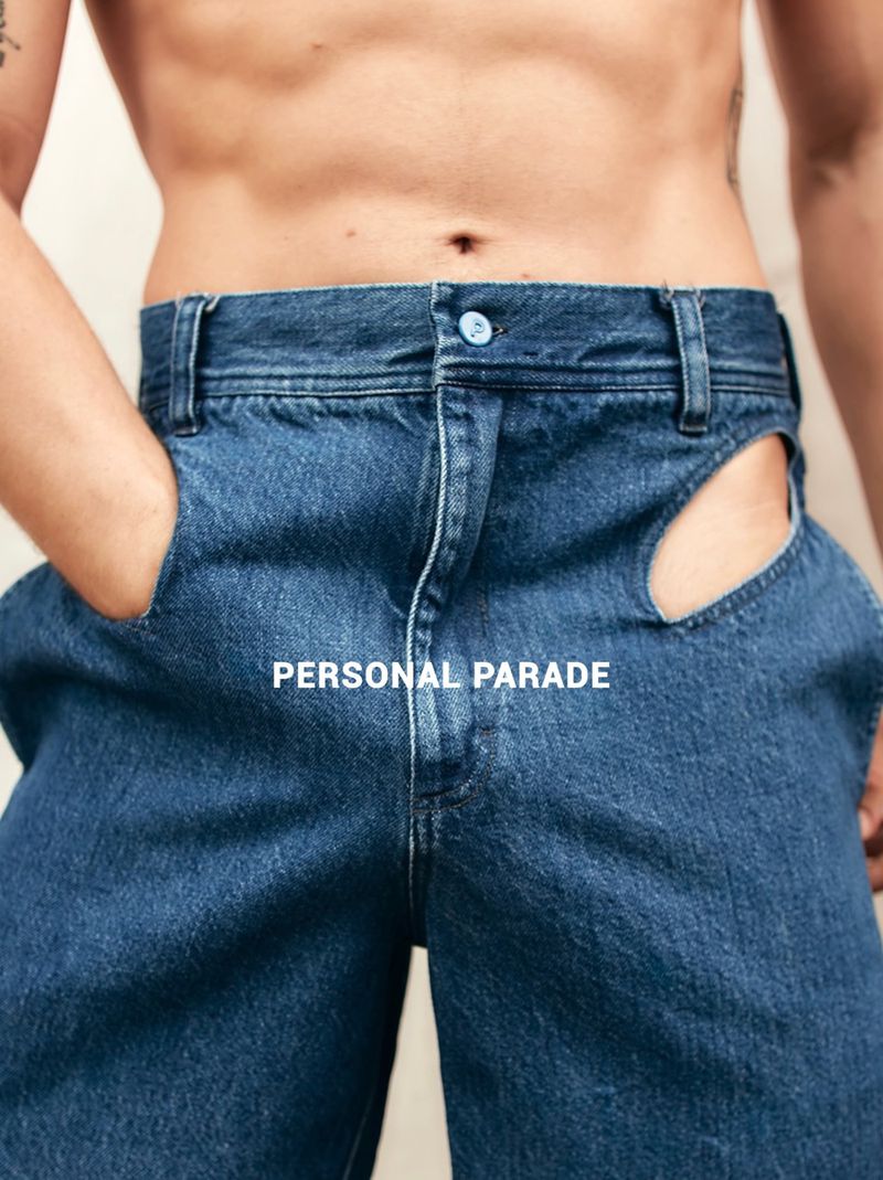 Denim jeans with hip cutouts on torso of a male model with hands in pockets against an off-white background in a studio setting.