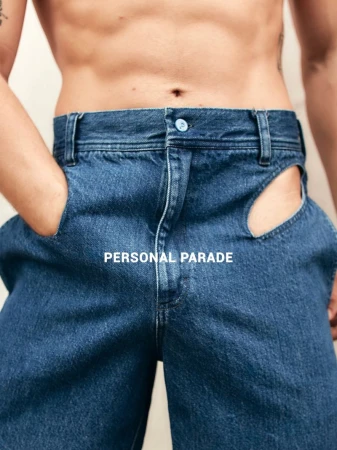 Denim jeans with hip cutouts on torso of a male model with hands in pockets against an off-white background in a studio setting.