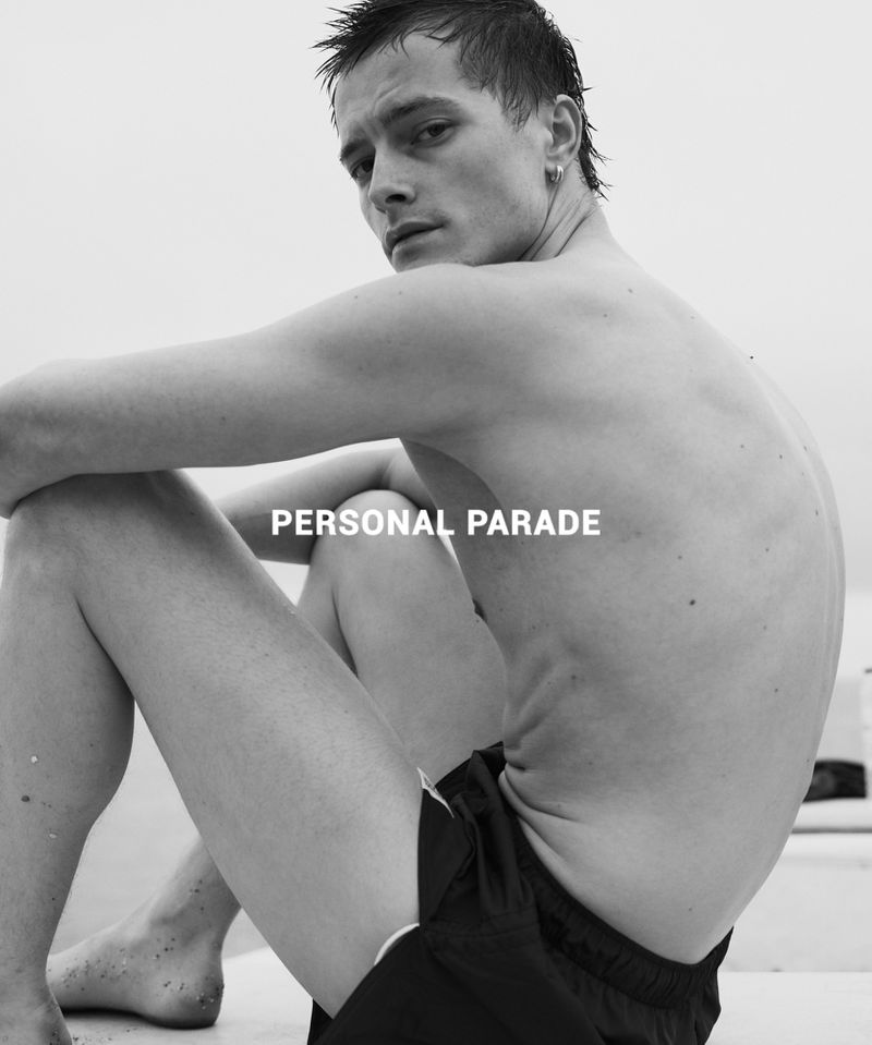 Black and white portrait of a young man sitting shirtless in swim trunks with his arms resting on his legs, looking at the camera.
