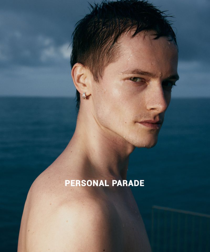A close-up studio shot captures a handsome adult male with short dark hair, glistening skin, and an earring, set against an ocean backdrop.