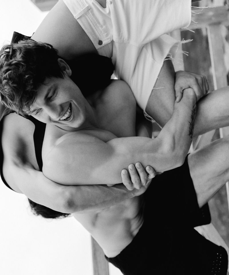 Monochrome portrait of two shirtless adult males embracing indoors, one smiling with strong musculature and holding the other.