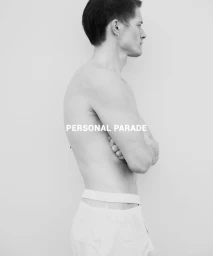 Black and white profile portrait of a shirtless male model with arms crossed wearing white underwear against a pale seamless background.