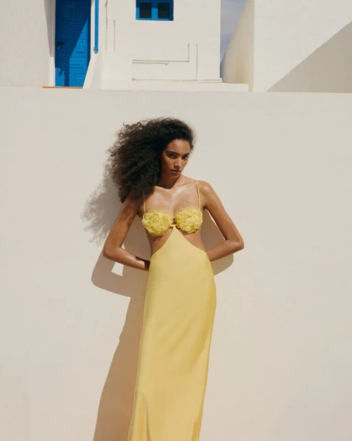 Elegant woman with dark curly hair poses in a yellow dress against a whitewashed wall and blue door backdrop on a sunny day.