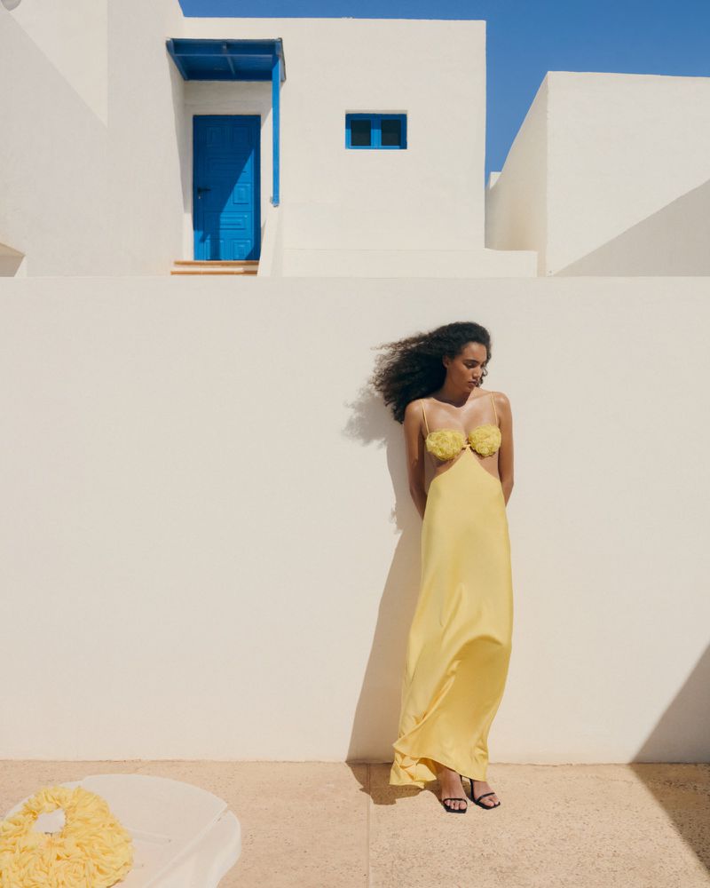 Woman in a yellow dress poses against a white wall on a sunny day. Building with blue door and window in background.
