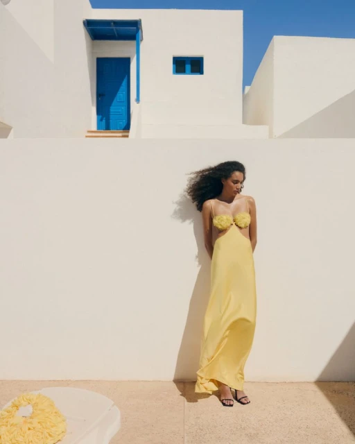Woman in a yellow dress poses against a white wall on a sunny day. Building with blue door and window in background.