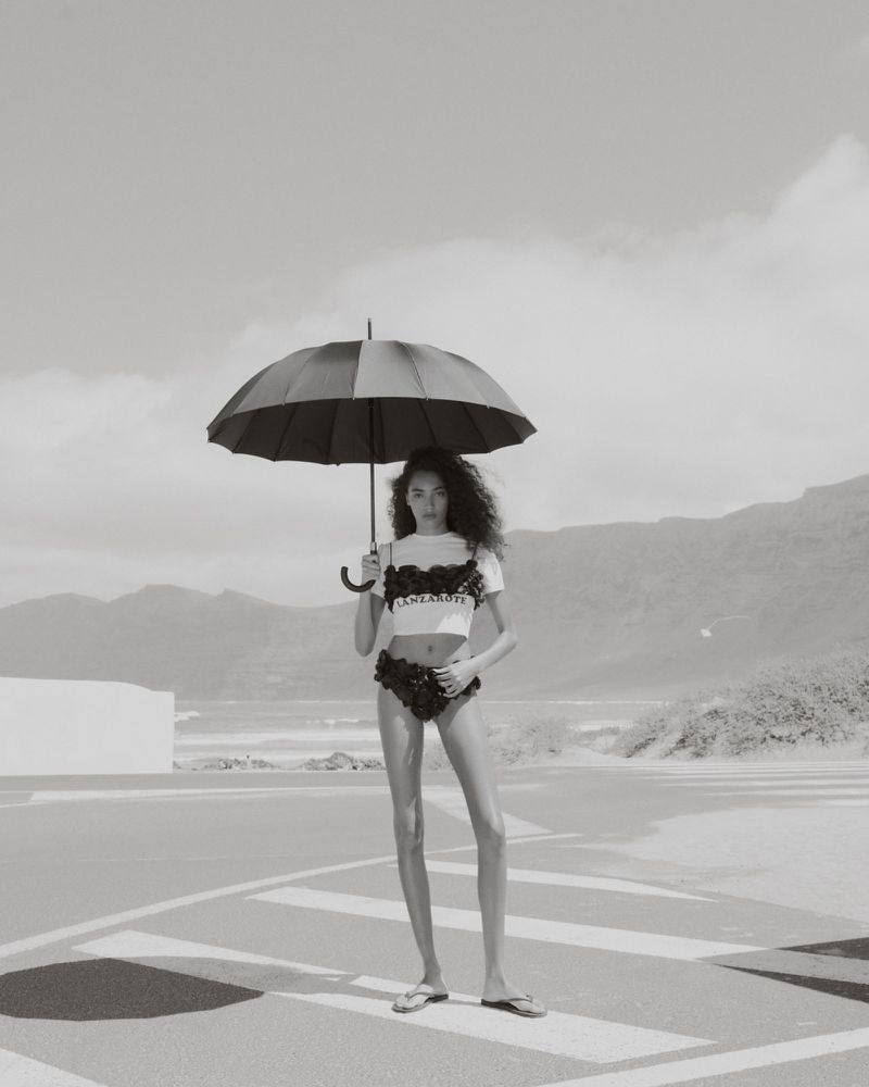 Monochrome portrait of a woman standing on a crosswalk, holding an umbrella, with mountains and a muted sky in the background.
