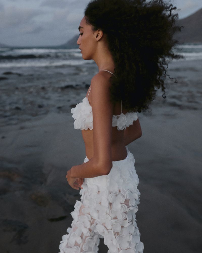 A woman in a white floral outfit stands on a beach, waves and cloudy sky in the background. Coastal fashion photoshoot.