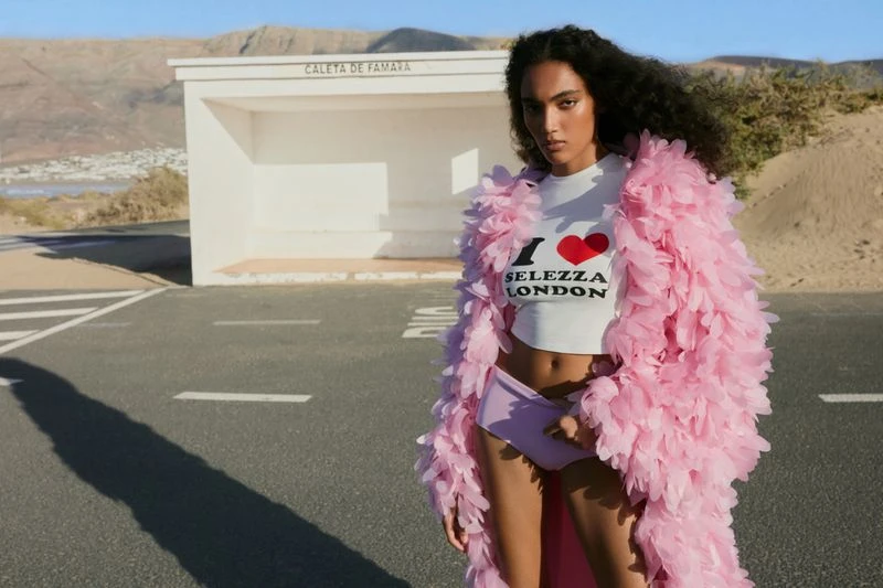 Fashionable woman in desert landscape: White top, lilac bikini bottom, and pink feathered coat. Clear blue sky and asphalt road.