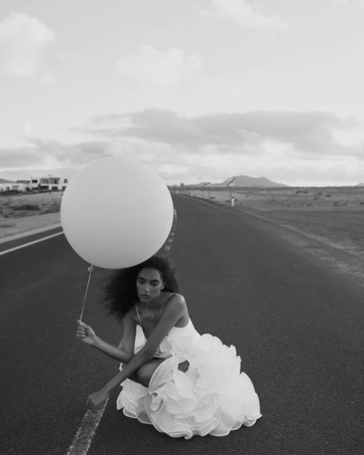 Person in white ruffled dress squats on asphalt road holding a large balloon, with flat landscape and cloudy sky in the background.