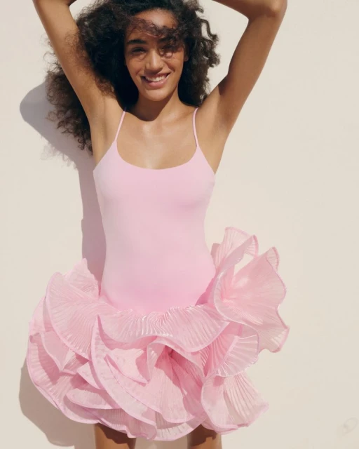 A smiling woman in a pink dress stands with arms raised against a neutral wall, bathed in sunlight and showcasing a blend of fashion and simplicity.