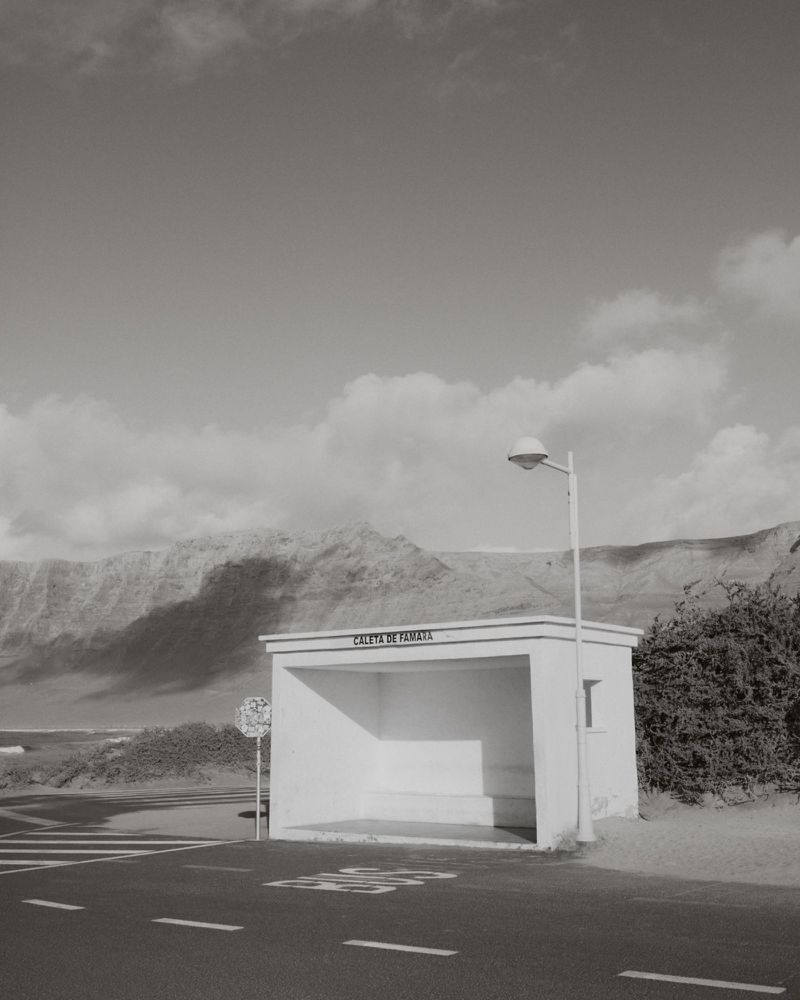Monochrome landscape featuring a white bus stop shelter with signage and mountains in the background under a cloudy sky.