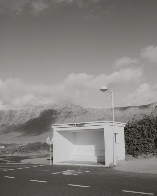 Monochrome landscape featuring a white bus stop shelter with signage and mountains in the background under a cloudy sky.