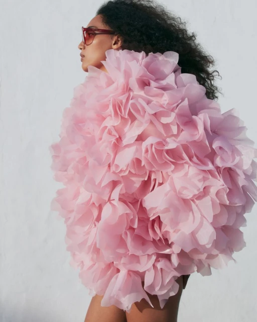 Fashion photo of an adult female wearing pink ruffled jacket and sunglasses posing against a white wall in bright sunny light.