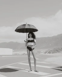 Model holding a black umbrella on a coastal road wearing a cropped top and ruffled briefs in black and white.